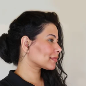 Wesliene Anália em uma pose de perfil com cabelos pretos ondulados, penteados em rabo de cavalo baixo, usando blusa social preta em fundo neutro